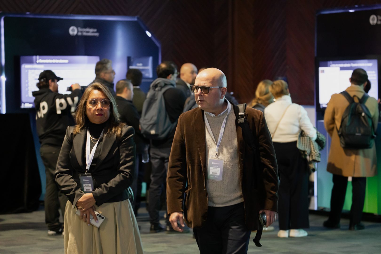 Dos asistentes al AI Academic Exchange caminan frente a los m&oacute;dulos interactivos donde se presentan proyectos de inteligencia artificial dentro del IA in Education Summit.