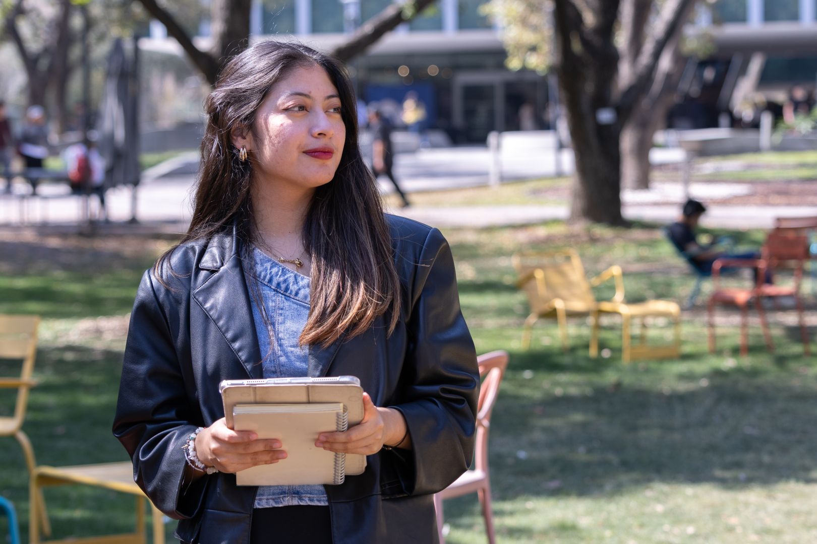 Jana, estudiante del Tec, sostiene una libreta y una tablet mientras camina por una zona verde del campus.