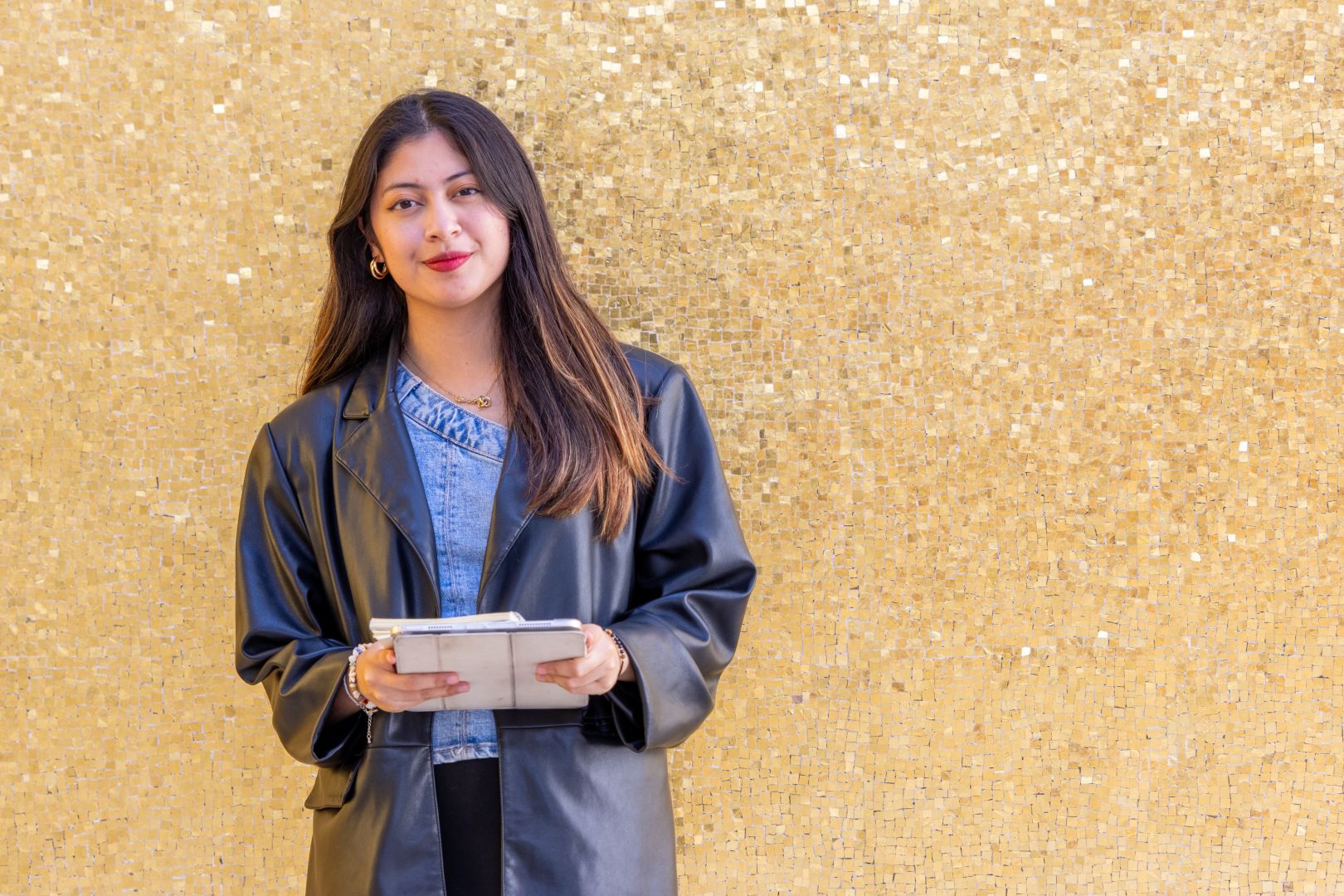 Retrato de Jana frente a un muro dorado, sosteniendo una tablet y mirando a la c&aacute;mara.