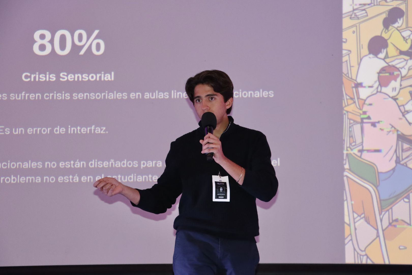 Estudiante de PrepaTec Morelia sostiene un micr&oacute;fono y expone el proyecto Elysium frente a una pantalla que muestra datos sobre crisis sensorial en aulas tradicionales. Detr&aacute;s se observan gr&aacute;ficos e ilustraciones relacionadas con el entorno escolar, durante su presentaci&oacute;n en el Encuentro Nacional de L&iacute;deres con Sentido Humano.