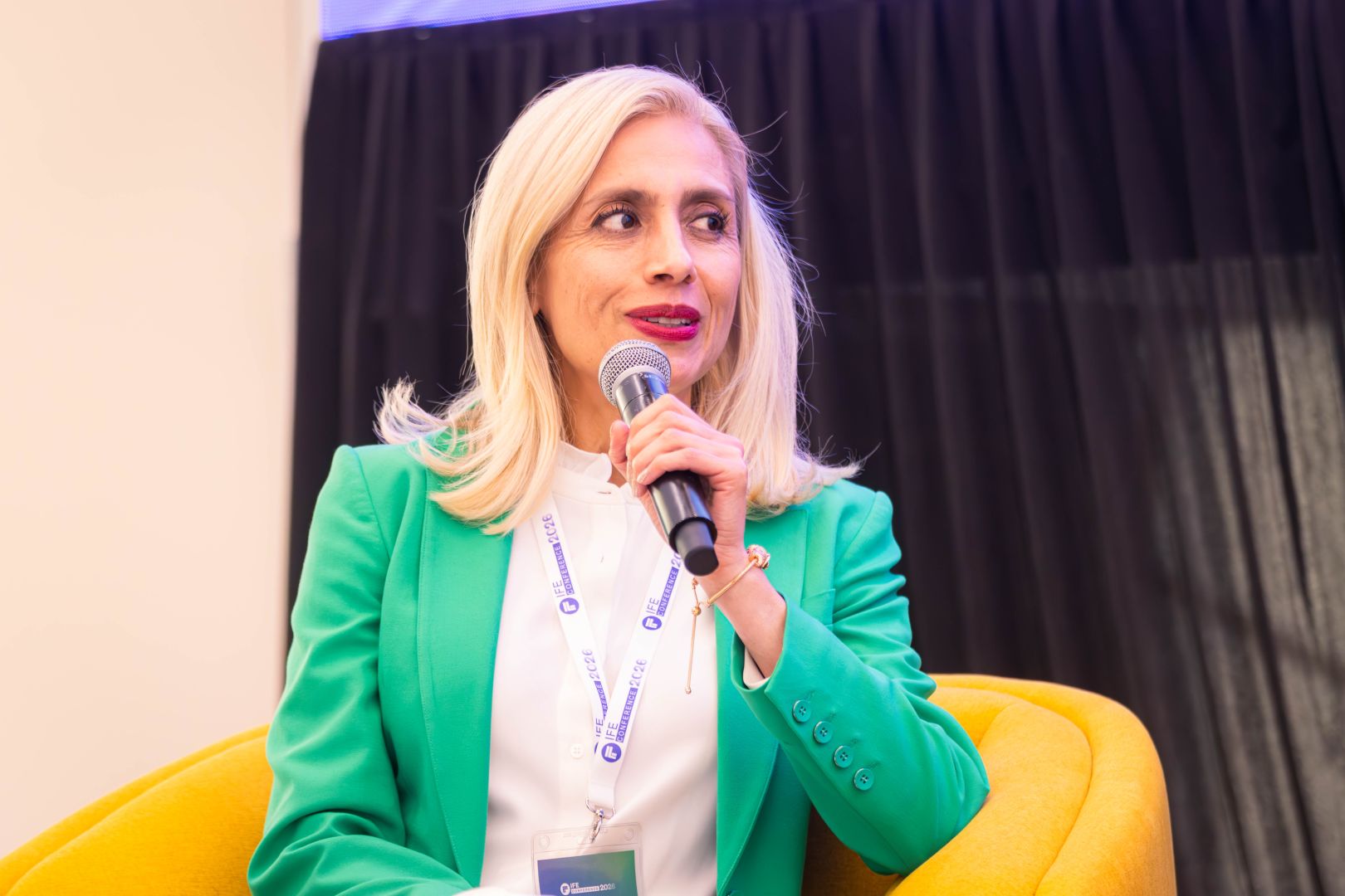 Ponente sentada en un sill&oacute;n amarillo, vestida con saco verde y blusa blanca, sostiene un micr&oacute;fono mientras participa en un panel sobre innovaci&oacute;n educativa frente a una audiencia.