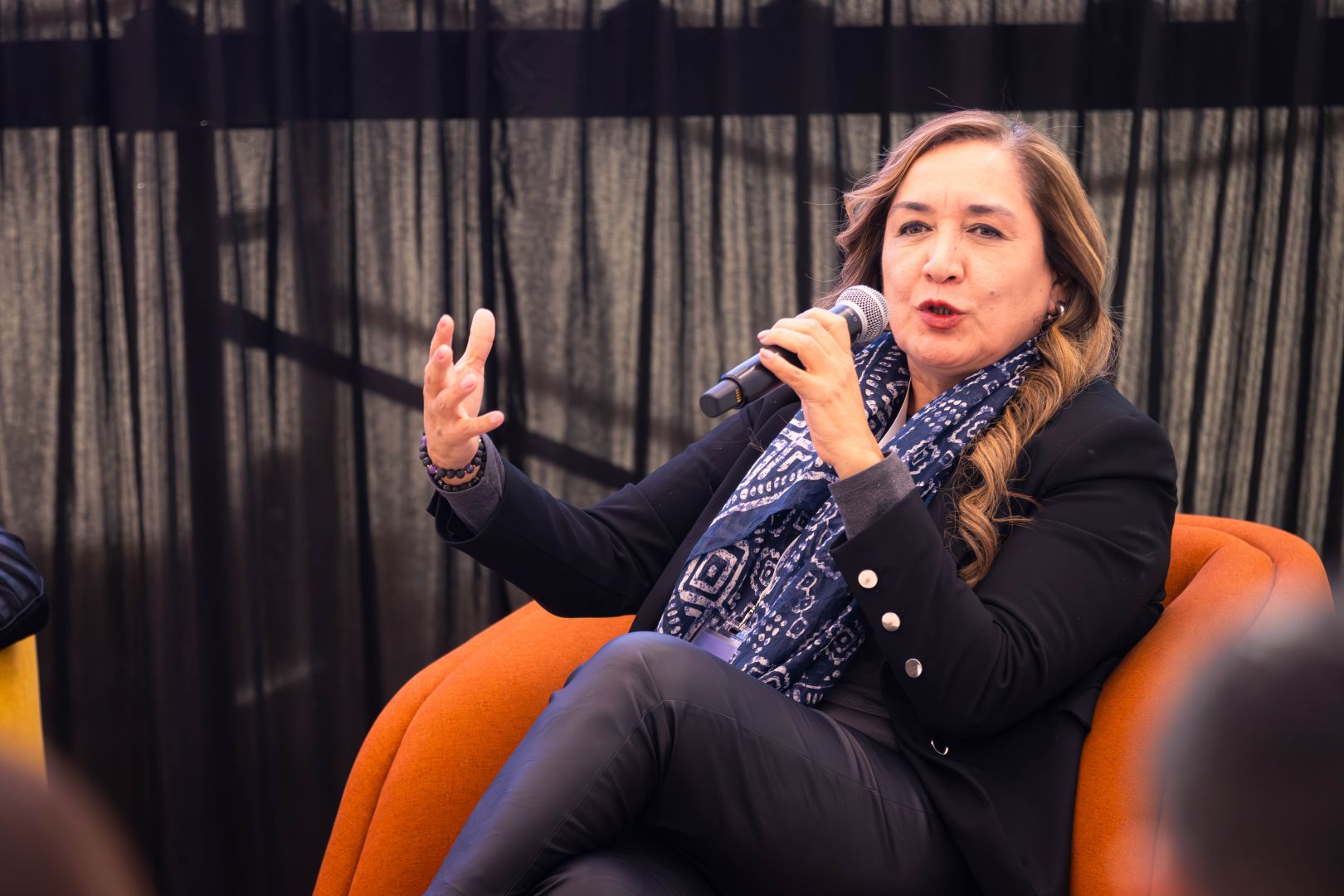 Ponente sentada en un sill&oacute;n naranja, con saco oscuro y pa&ntilde;uelo, habla al micr&oacute;fono mientras gesticula durante un panel frente a asistentes.