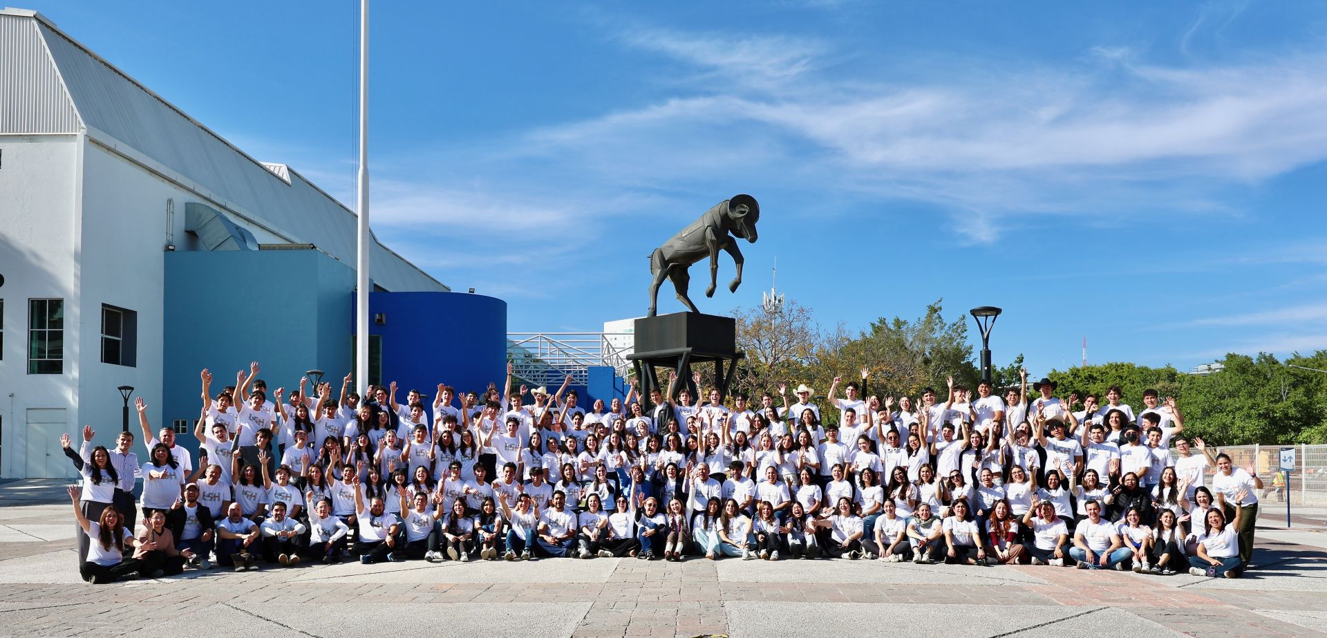 Fotograf&iacute;a panor&aacute;mica de un gran grupo de estudiantes y profesores de PrepaTec vestidos con playeras blancas, reunidos al aire libre en un campus universitario. Las y los participantes levantan las manos y sonr&iacute;en frente a una escultura emblem&aacute;tica, reflejando un ambiente de celebraci&oacute;n, comunidad y participaci&oacute;n colectiva.