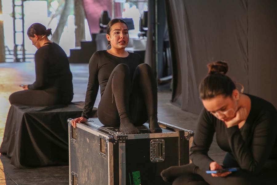 Tres jóvenes vestidas de negro sentadas sobre cajas negras en un escenario. 