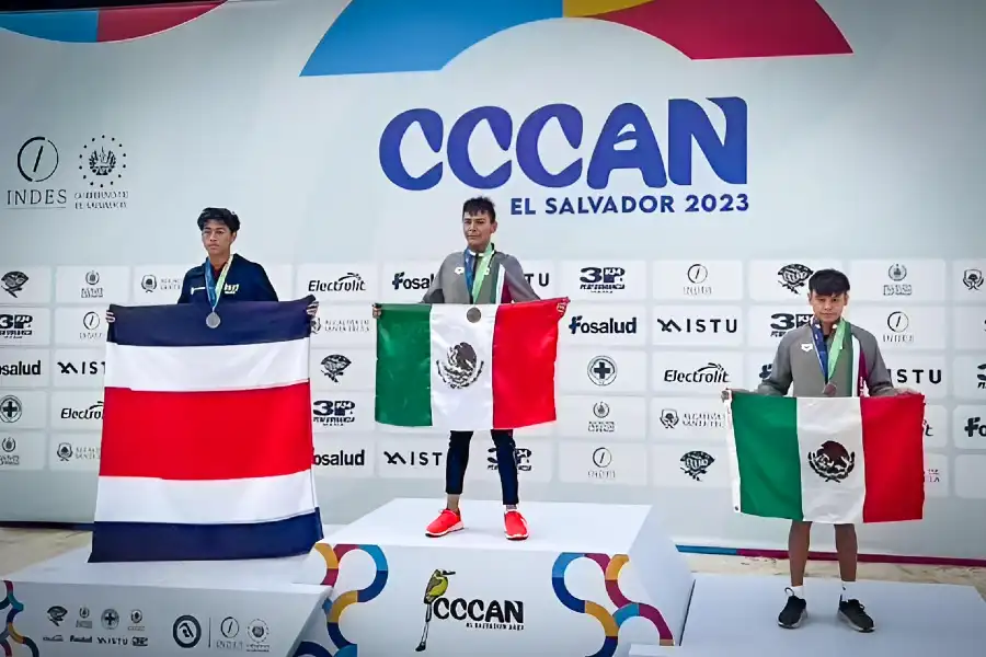 Diego Lizardi, nadador hidalguense, en el podio con la medalla de oro en el CCCAN El Salvador 2023, sosteniendo la bandera de México junto a los medallistas de Costa Rica y México.