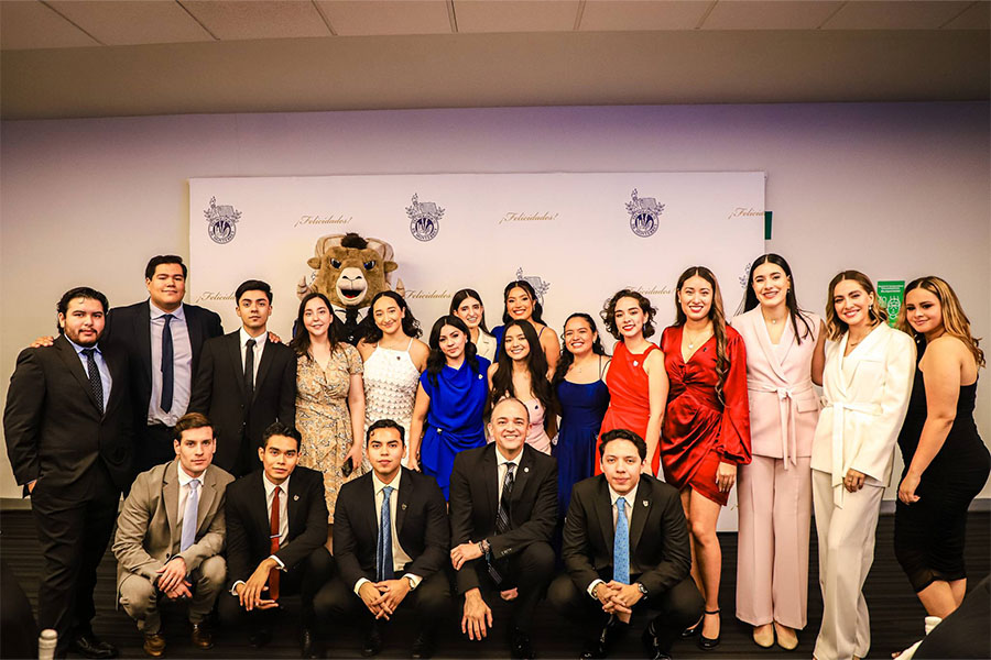 Generación de graduados y graduadas, con sus ganadoras al Premio Ceneval, posan para una fotografía en el día de su graduación con un fondo blanco que muestra el escudo del Tec de Monterrey y la palabra felicidad