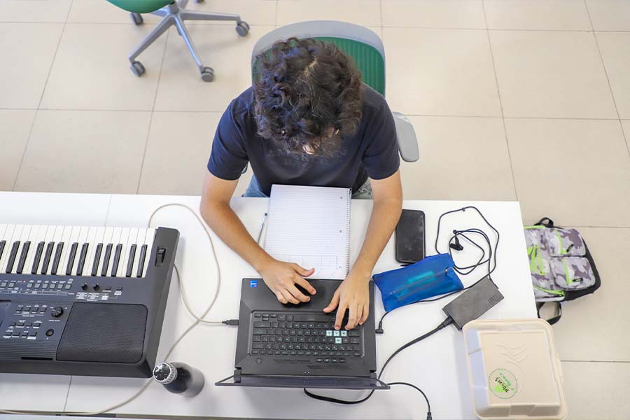 Vista en picada de un joven tecleando en una computadora con un teclado musical al lado. 