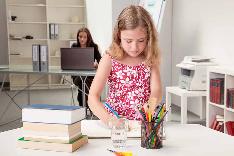 Niña realizando actividades, al fondo se observa su mamá en un escritorio trabajando