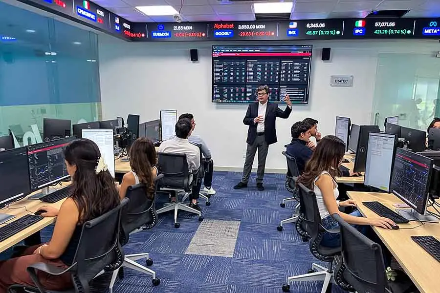 Profesor al frente de un salón, con una pantalla que muestra muchos números y datos financieros. Frente a el están sentados en dos filas alumnos en estaciones de cómputo con pantallas que muestran los mismos números.