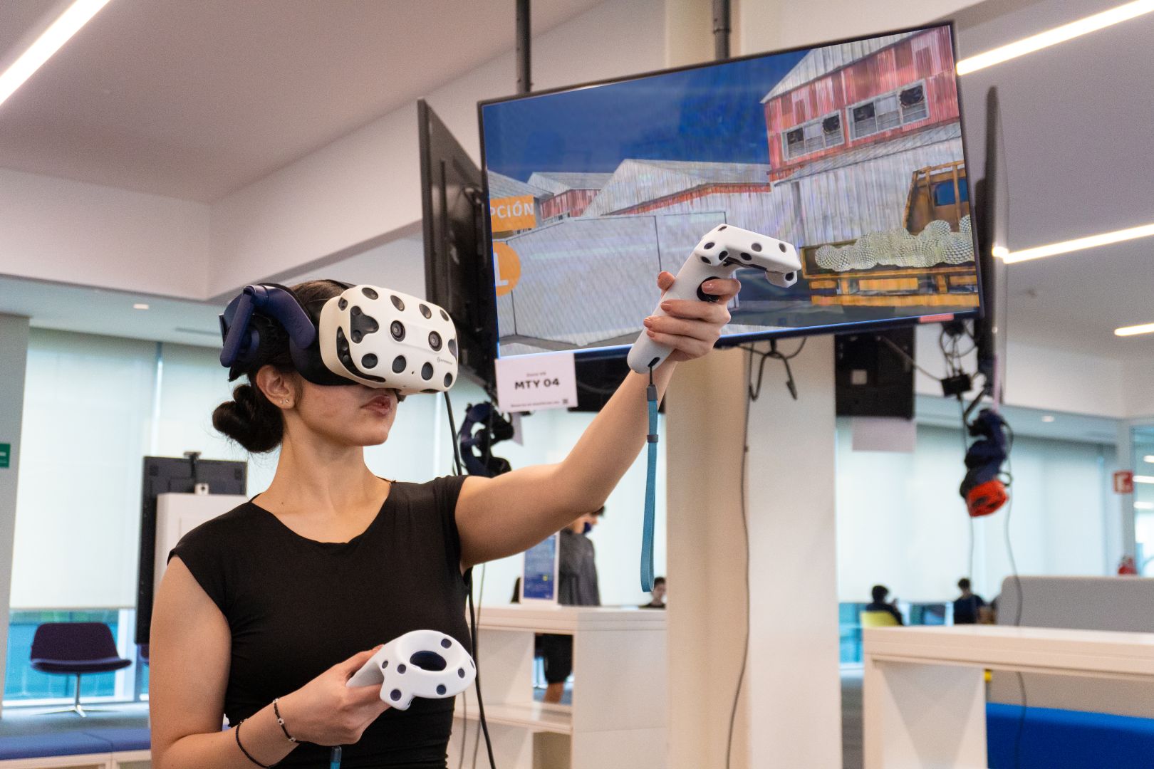 Estudiante utilizando visor y controles de realidad virtual para interactuar con una simulación de una planta tequilera proyectada en una pantalla dentro de un espacio de aprendizaje del Tec.
