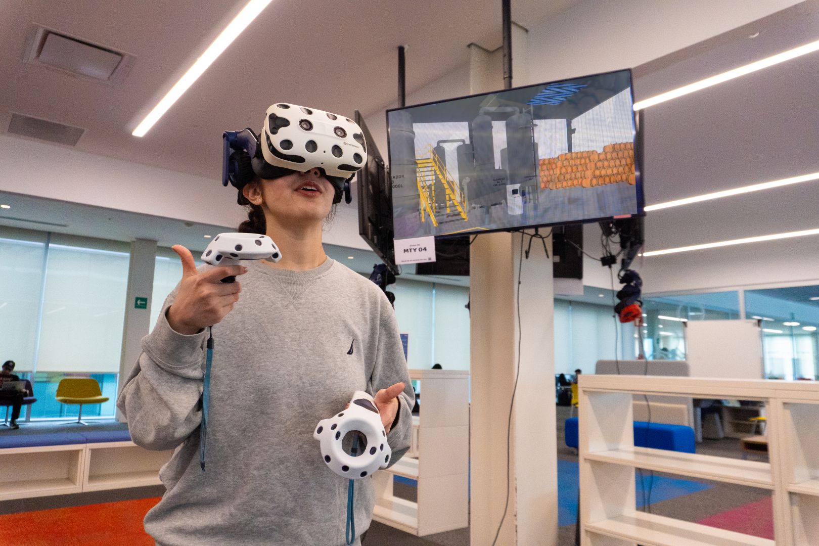 Estudiante utilizando visor y controles de realidad virtual mientras observa en una pantalla la simulaci&oacute;n de una l&iacute;nea de producci&oacute;n de tequila en un entorno virtual.