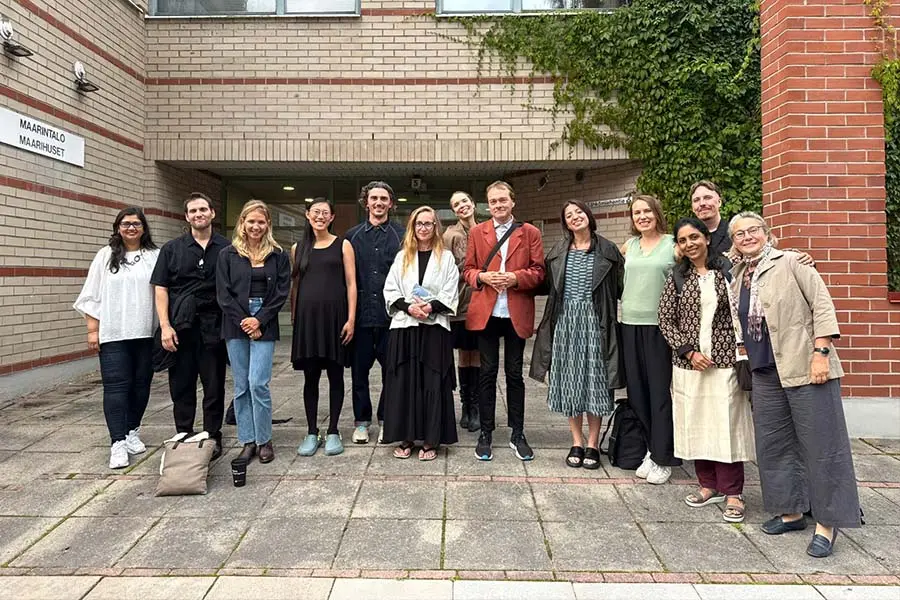 13 profesores de arquitectura posan en grupo frente a edificio de ladrillos. 