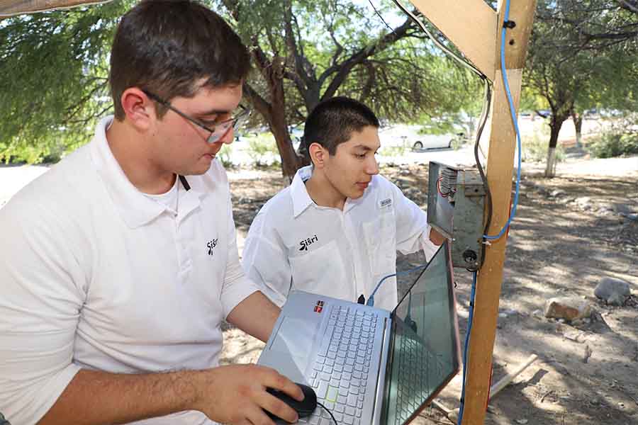 Un alumno revisa la caja de comando de un sistema de riego mientras que otro sostiene una computadora.