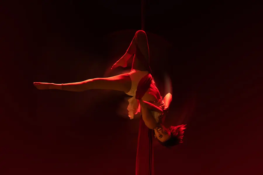 Mujer joven en el aire de cabeza con una pierna extendida horizontalmente, sostenida por una tela roja enredada en su cuerpo, iluminada por luz roja con un fondo negro.
