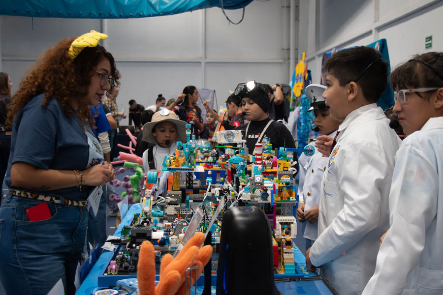 Niños utilizando bata exponen proyecto de FIRST LEGO League frente a presentadora en PrepaTec Laguna