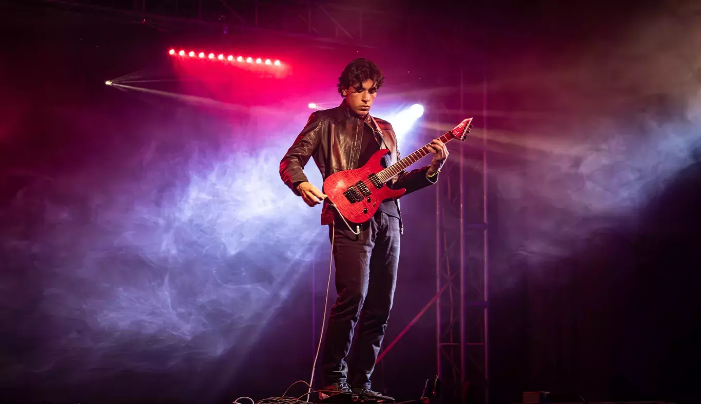 Andrés, guitarrista del ensamble de músicos de Arte y Cultura, posando en el escenario con su guitarra roja bajo una atmósfera de luces y humo.