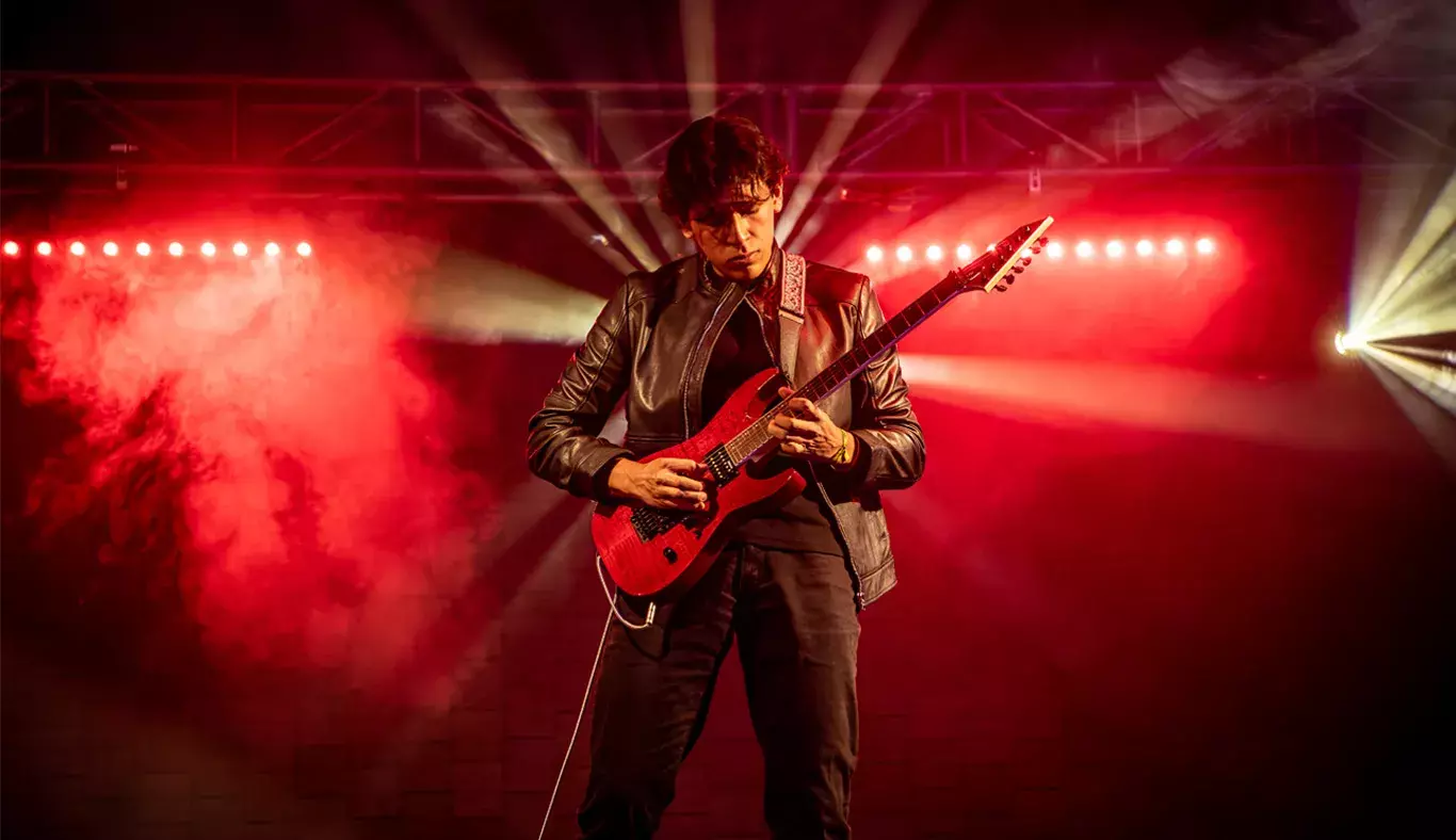 Andrés Ortega en un solo de guitarra, rodeado de luces rojas y un fondo de humo.