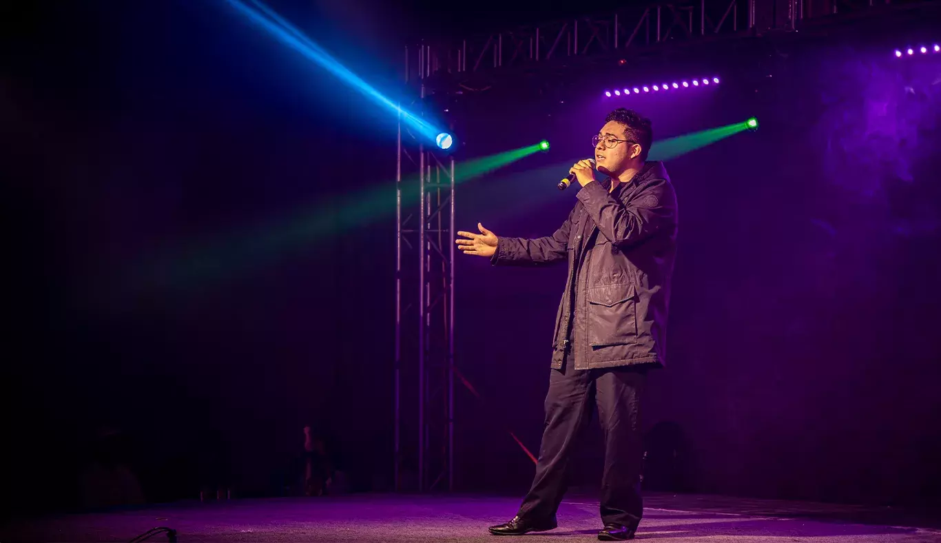 Eric González, estudiante de ingeniería y cantante, interpretando en el escenario. Con micrófono en mano y luces de colores que lo iluminan.