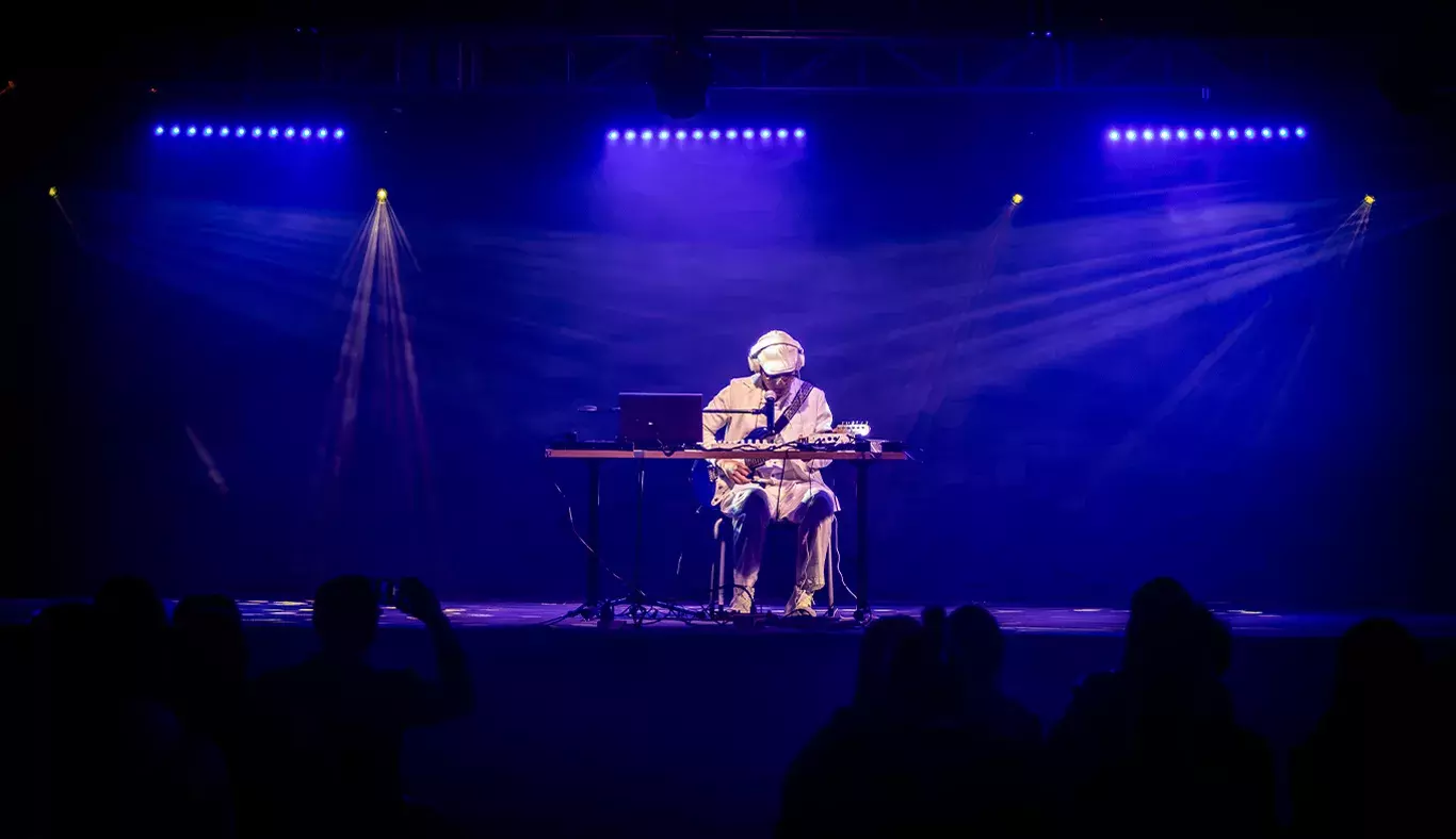 Jesús Alejandro, estudiante y músico, tocando en un escenario iluminado en tonos azules durante su presentación. Sentado con su equipo, la audiencia observando su interpretación.