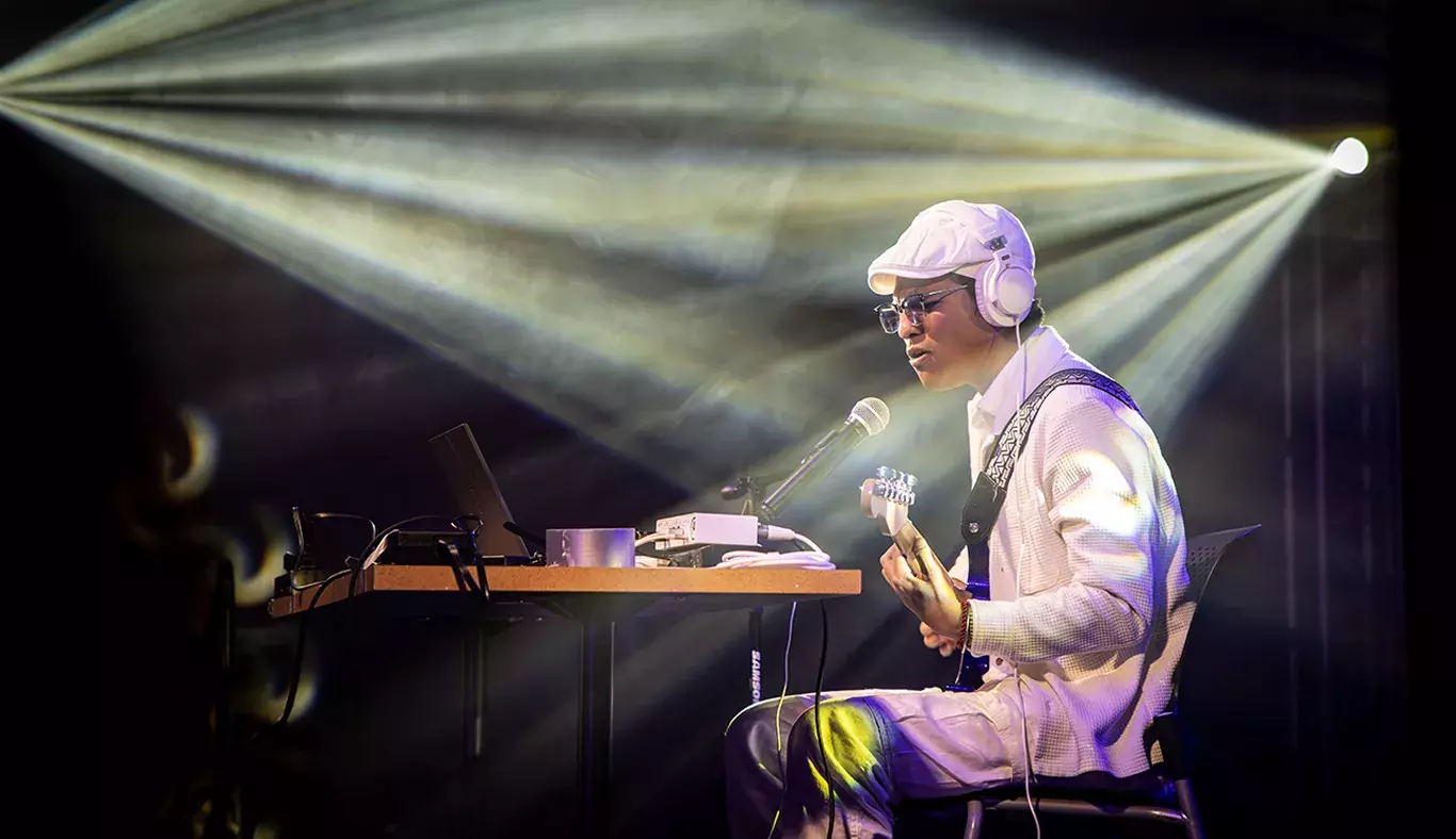 Jesús Alejandro, músico especializado en looping, en plena actuación. Sentado frente a su equipo de sonido, usa audífonos y toca su guitarra, mientras las luces crean un ambiente en el escenario.