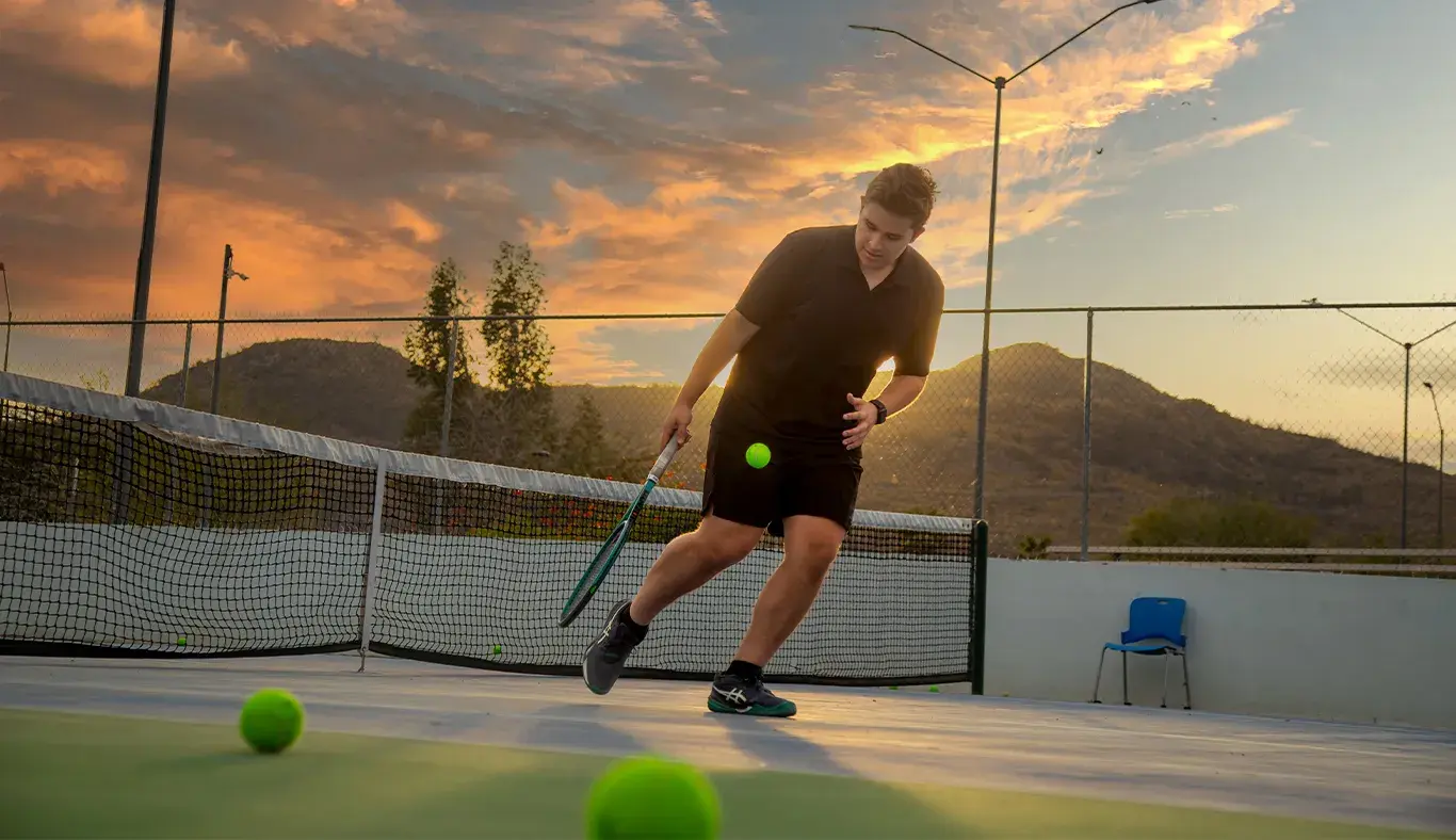 Joven universitario jugando tenis con atardecer de fondo.