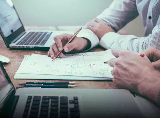 Toma cerrada de las manos de dos personas escribiendo sobre un papel con laptops a los lados. 