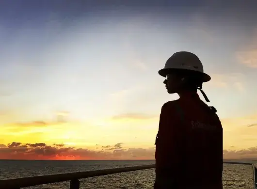 Ashly Hinojos con casco y uniforme naranja mirando el atardecer sobre el mar desde una plataforma petrolera.