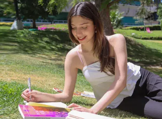Alumna escribiendo al aire libre con libros a su alrededor