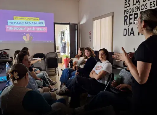 Mujer dando una platica frente a un grupo de mujeres jóvenes.