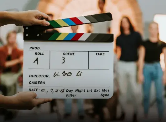 Primer plano de una claqueta de cine, al fondo se ve un grupo de personas desenfocadas.