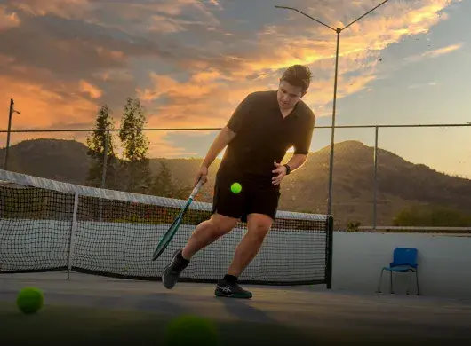 Joven universitario jugando tenis con atardecer de fondo.