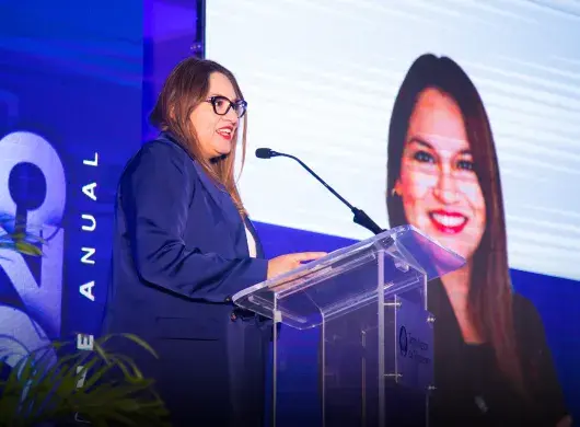 Mujer hablando en un podio frente a pantallas. 