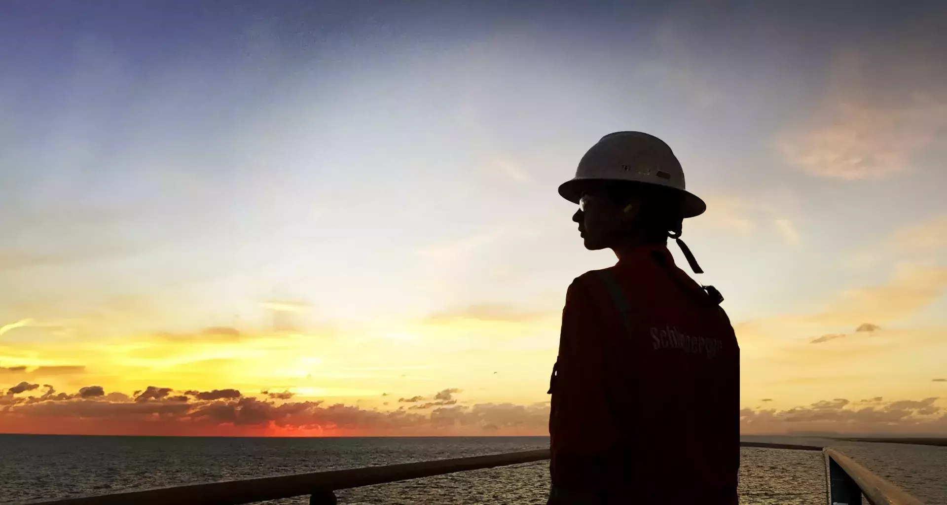 Ashly Hinojos con casco y uniforme naranja mirando el atardecer sobre el mar desde una plataforma petrolera.