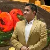 Profesor de arte usa traje color café y de fondo tiene una de sus pinturas en óleo con tomates y chiles