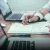 Toma cerrada de las manos de dos personas escribiendo sobre un papel con laptops a los lados. 