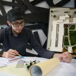 Jóven sonriente trabajando en una mesa con material de arquitectura alrededor 