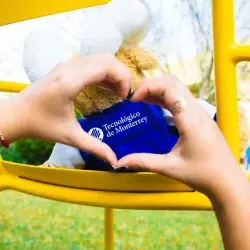 Peluche con chamarra del Tec de Monterrey tras la figura de un corazón formado por manos