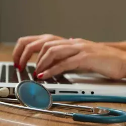 Una mujer escribiendo sobre el teclado de una computadora, a su lado tiene un estetoscopio