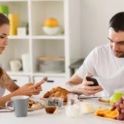 Dos personas comiendo mientras revisan sus dispositivos móviles.