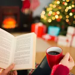 Mano de una mujer joven, sosteniendo un libro, y se observa al fondo el pino de navidad encendido