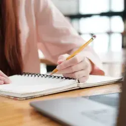Mujer escribe en su libreta mientras observa su computadora portátil.
