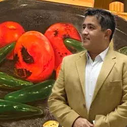 Profesor de arte usa traje color café y de fondo tiene una de sus pinturas en óleo con tomates y chiles