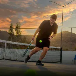 Joven universitario jugando tenis con atardecer de fondo.