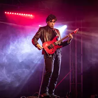 Andrés, guitarrista del ensamble de músicos de Arte y Cultura, posando en el escenario con su guitarra roja bajo una atmósfera de luces y humo.