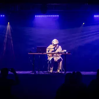 Jesús Alejandro, estudiante y músico, tocando en un escenario iluminado en tonos azules durante su presentación. Sentado con su equipo, la audiencia observando su interpretación.