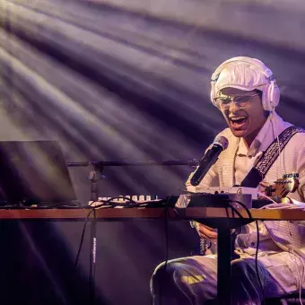 Jesús Alejandro, guitarrista y cantante, expresando emoción mientras canta en el escenario de Tec's Got Talent. Rodeado de su equipo de música, utiliza audífonos y micrófono, bajo los haces de luz.