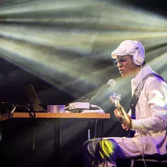 Jesús Alejandro, músico especializado en looping, en plena actuación. Sentado frente a su equipo de sonido, usa audífonos y toca su guitarra, mientras las luces crean un ambiente en el escenario.