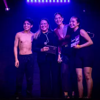 Nallely y Karol, ganadores del segundo lugar en Tec's Got Talent, posando junto a las mentoras de la comunidad KREI, sonriendo y sosteniendo su premio, bajo una iluminación azul.