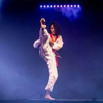 Rod Mendoza, estudiante y artista marcil, ejecutando una patada en su presentación, vestido con su uniforme de y rodeado de luces.