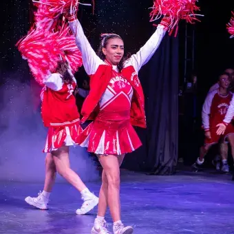 Estudiante caracterizada como porrista del equipo Wildcats realiza una coreografía con pompones durante el musical High School Musical: On Stage, en el escenario del campus Hidalgo.